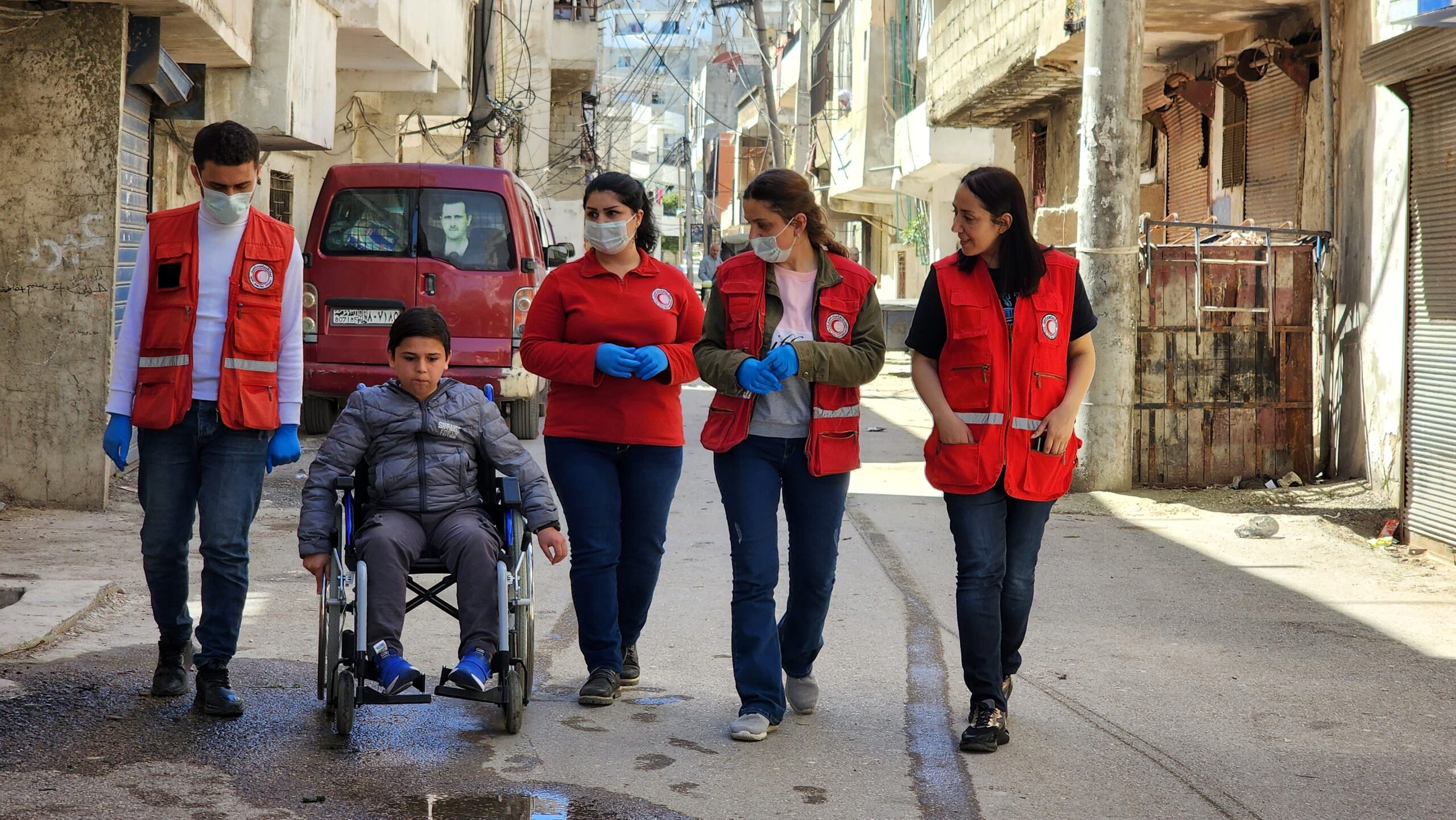 Red Cross workers wearing red vests and masks provide assistance to a young boy in a wheelchair on a damaged street lined with dilapidated buildings and exposed wires, illustrating humanitarian relief efforts after the 2023 earthquake in Syria